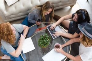 4 Frauen sitzen um einen Glastisch mit einem Laptop darauf.