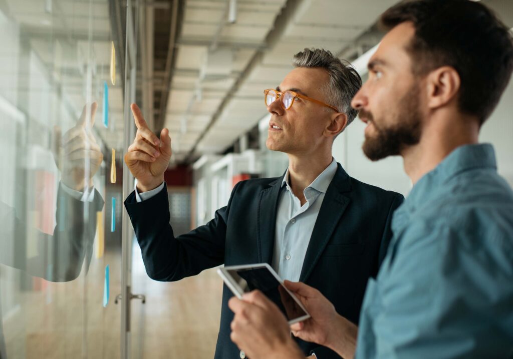 Zwei Männer stehen an einer Glaswand mit Haftnotizen und diskutieren mit Brainymotion über Ideen. Der eine zeigt auf die Notizen, der andere hält ein digitales Tablet. Beide arbeiten konzentriert und engagiert in einer modernen Büroumgebung.