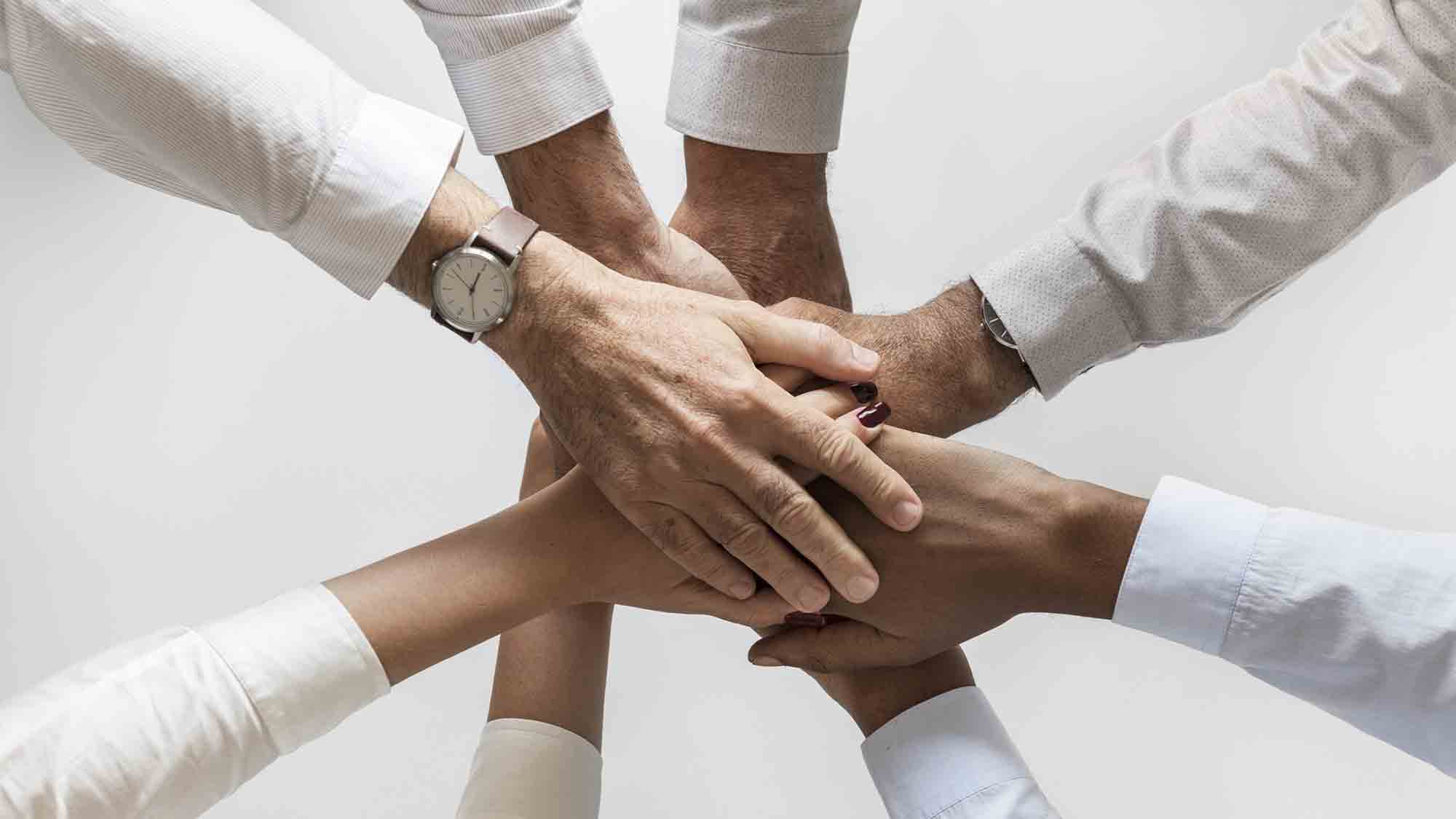 Sechs Hände unterschiedlicher Individuen, die in der Mitte einen einheitlichen Stapel bilden. Jede Person trägt ein anderes langärmeliges Hemd, was Teamwork, Einheit und Zusammenarbeit suggeriert. Eine Person trägt eine Armbanduhr. Der Hintergrund ist schlicht weiß.