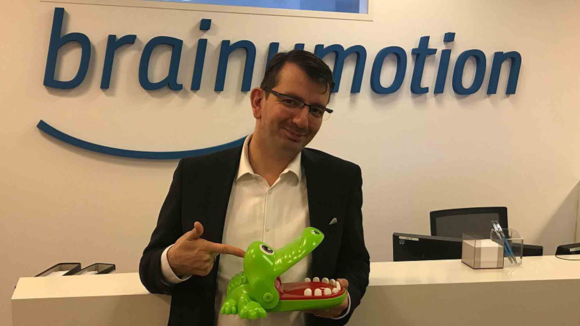 Eine Person mit Brille und dunklem Blazer steht vor einem Schild mit der Aufschrift „brainymotion“. Sie hält einen grünen Spielzeugalligator in der Hand und zeigt lächelnd darauf. Im Hintergrund sind ein Empfangstresen und Büromaterial zu sehen.