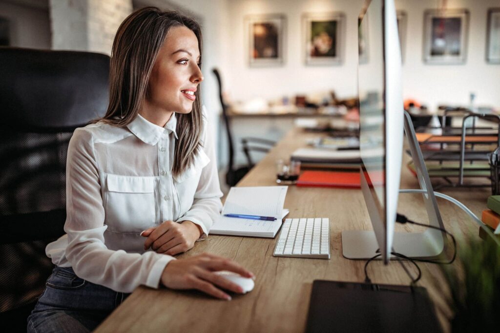 Eine Frau sitzt an einem Schreibtisch in einem Brainymotion-Büro, benutzt eine Computermaus und schaut auf einen Monitor. Auf dem Schreibtisch liegen ein Notizbuch, ein Stift und eine Tastatur. Im Hintergrund befinden sich Büroartikel und gerahmte Bilder.