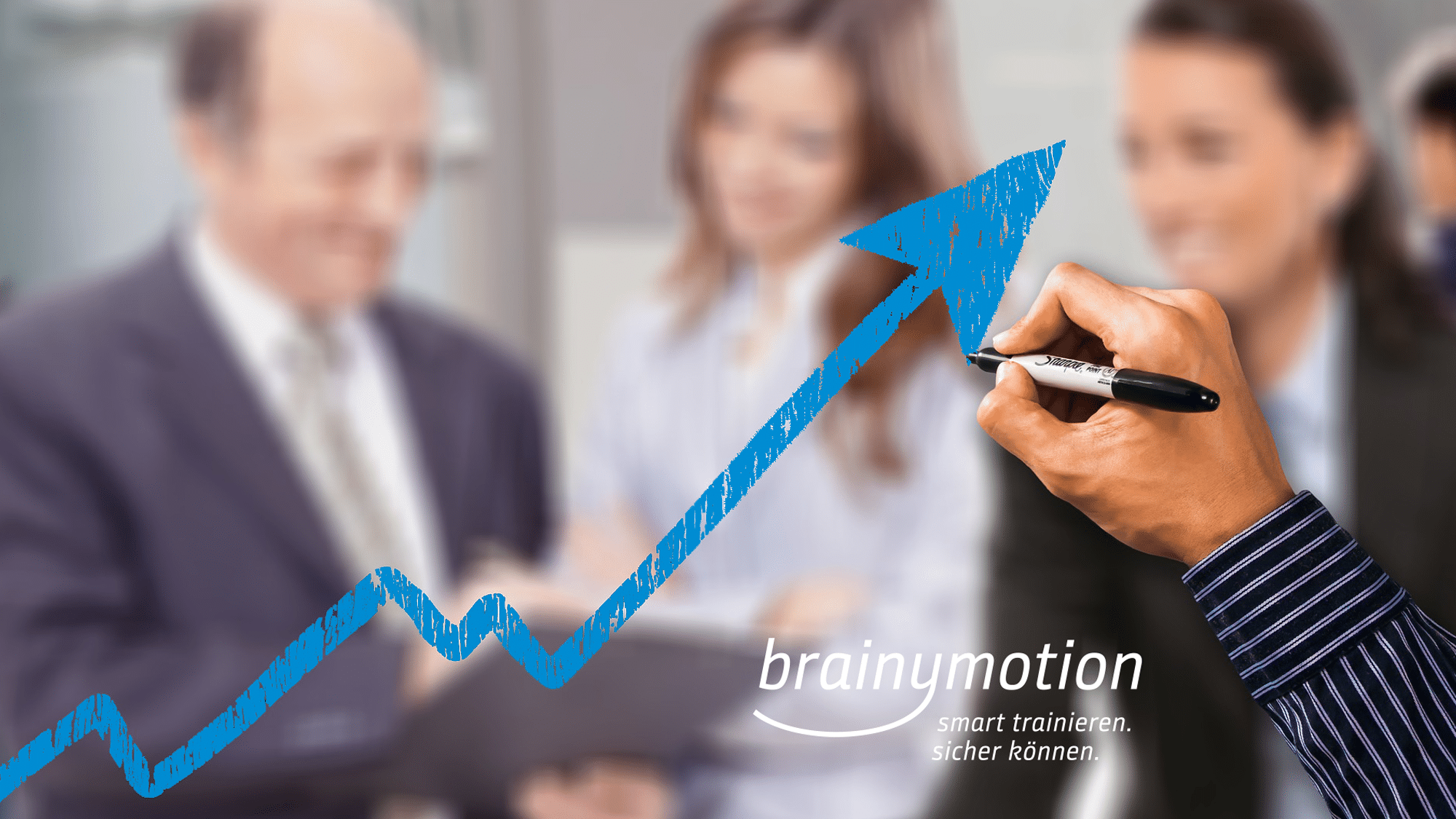 Eine Person im Business-Anzug malt mit schwarzem Marker einen blauen, nach oben weisenden Pfeil auf Glas. Im Hintergrund sind zwei weitere Personen in Business-Kleidung unscharf zu erkennen. Zu sehen ist der Text „brainymotion smart trainieren. sicher können.“