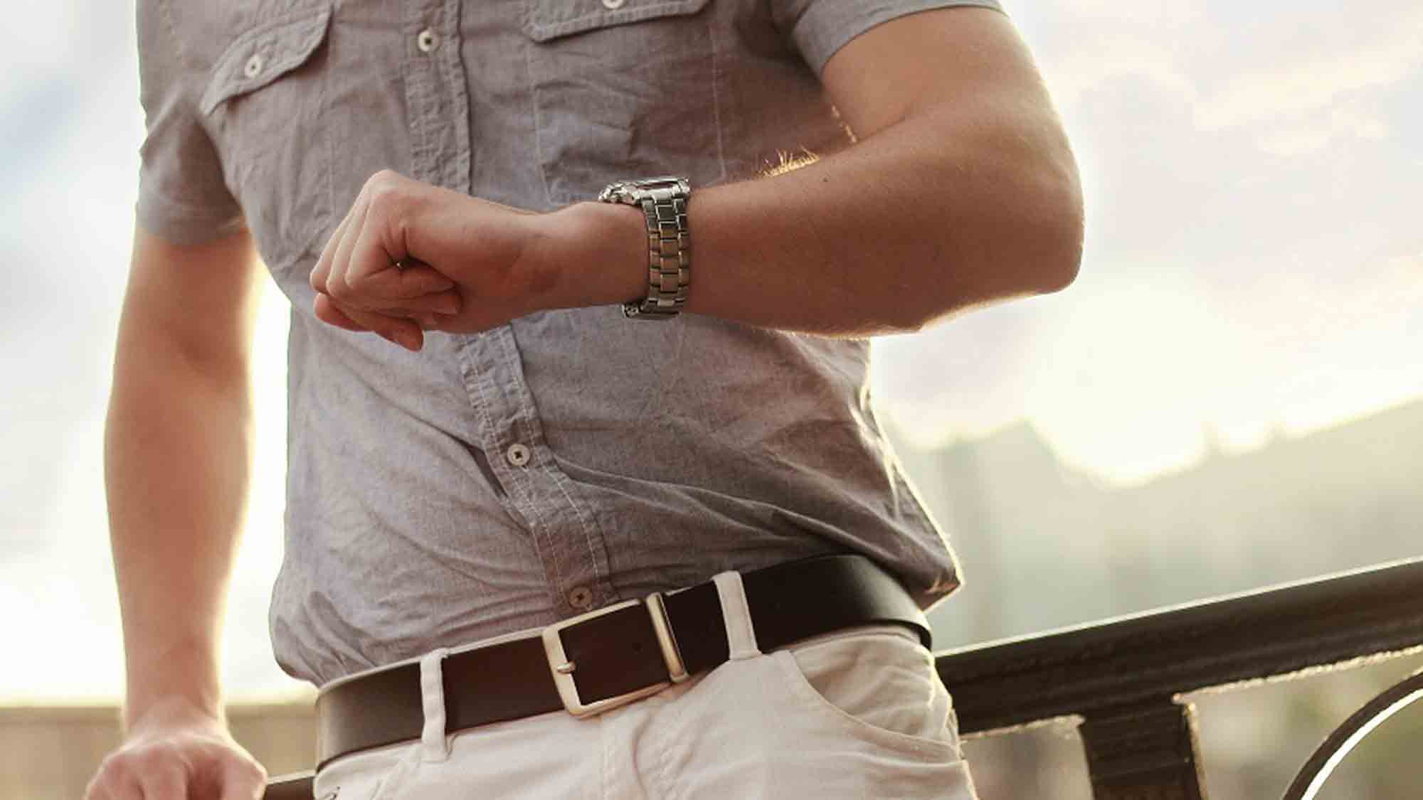 Eine Person in einem grauen Button-Down-Hemd und weißen Hosen schaut auf eine Armbanduhr. Das Bild ist so zugeschnitten, dass Oberkörper und Arme zu sehen sind. Die Person steht an einem sonnigen Tag im Freien neben einem Geländer.