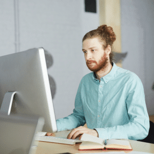 Eine Person mit langen, zu einem Dutt gebundenen Haaren und Bart arbeitet an einem Schreibtisch und blickt konzentriert auf einen großen Computerbildschirm. Sie trägt ein hellblaues Hemd mit Knöpfen und auf dem Schreibtisch liegt ein offenes Notizbuch. Der Hintergrund ist unscharf und lässt auf eine Büroumgebung schließen.