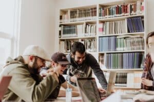 Eine Gruppe von vier Personen sitzt um einen Tisch herum, lächelt und schaut auf einen Laptop-Bildschirm. Sie scheinen sich in einer Bibliothek oder einem Arbeitszimmer zu befinden, im Hintergrund stehen Regale voller Bücher und Ordner. Die Atmosphäre ist lebhaft und kooperativ.