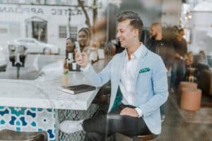 Ein junger Mann in einem hellblauen Blazer sitzt an einer schicken Café-Bar, lächelt und schaut auf sein Handy. Neben ihm auf der Theke steht ein Getränk, dazu ein geschlossenes Notizbuch. Im Hintergrund sind durch das Glas drei Personen zu sehen.