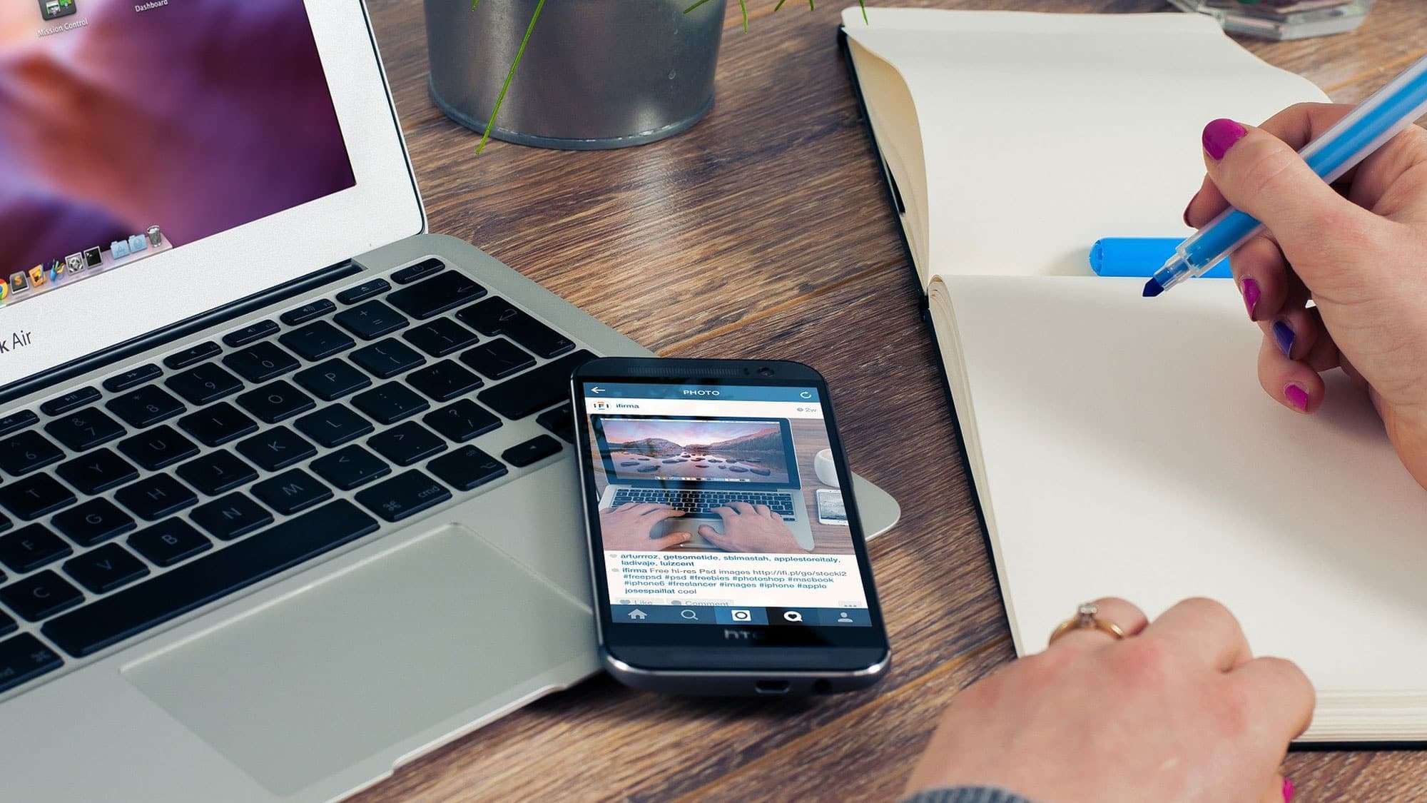 Eine Person arbeitet an einem Holzschreibtisch mit einem Laptop, einem Smartphone und einem Notizbuch. Auf dem Smartphone ist ein Bild von Händen zu sehen, die auf einem Computer tippen. Die Person schreibt mit einem blauen Stift in das Notizbuch. Im Hintergrund sind einige Pflanzenblätter zu sehen.