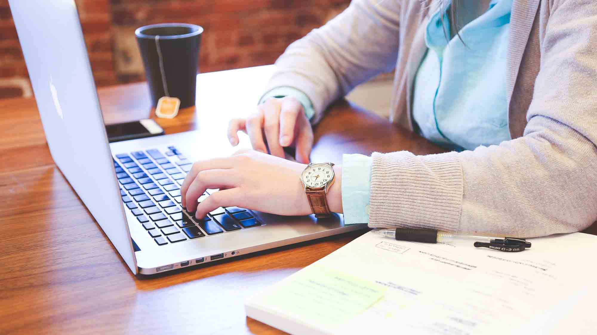 Eine Person tippt auf einem Laptop an einem Holzschreibtisch mit einer Tasse Tee, einem offenen Notizbuch und einigen Dokumenten. Sie trägt eine Uhr und ein hellblaues Hemd mit einem beigen Pullover. Die Umgebung scheint ein ungezwungener Arbeitsplatz zu sein.