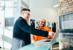 Zwei Männer unterhalten sich in einem scheinbar modernen Büro- oder Empfangsbereich. Ein Mann, der einen schwarzen Blazer trägt, zeigt auf einen Laptop-Bildschirm, während der andere Mann, der ein orangefarbenes Hemd trägt, lächelt und zuhört.