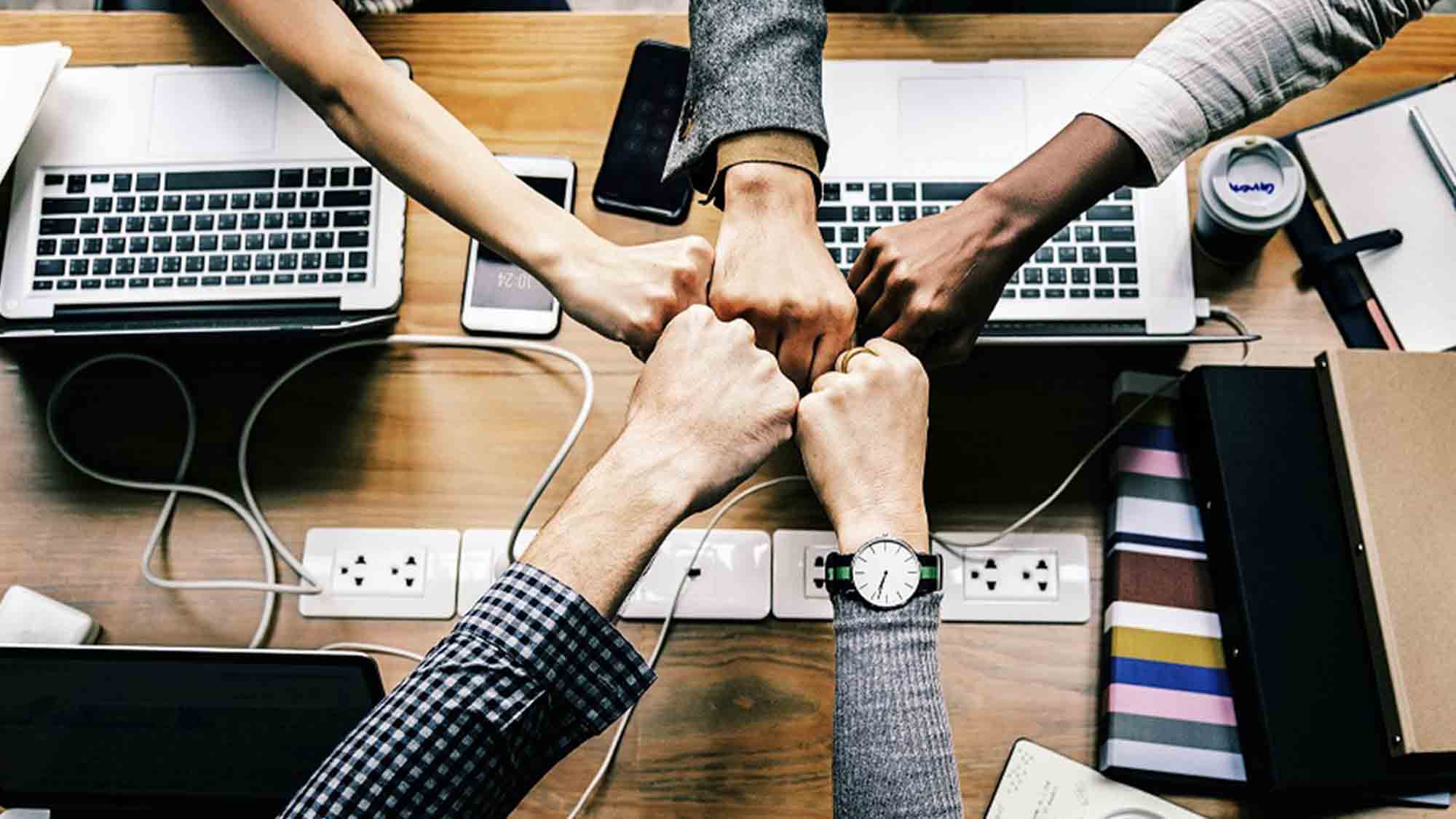 Fünf Personen stoßen über einem Holztisch mit Laptops, Notizbüchern und Telefonen mit den Fäusten zusammen. Ihre Hände treffen sich in der Mitte und symbolisieren Teamwork und Einheit. Auf dem Tisch sind mehrere Steckdosen zu sehen.