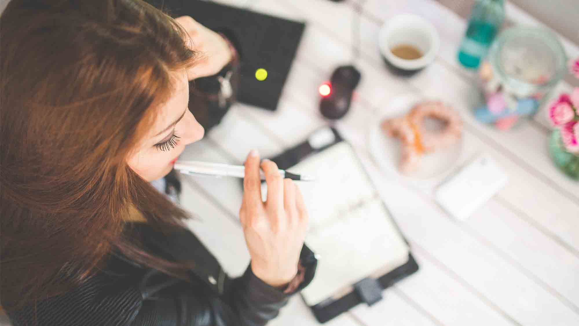 Eine Frau mit langen Haaren sitzt an einem Schreibtisch, hält einen Stift an die Lippen und schaut auf ein offenes Notizbuch oder einen Planer. Auf dem Schreibtisch liegen ein Laptop, eine Computermaus, eine Tasse Kaffee und etwas Gebäck. Sie scheint in Gedanken versunken zu sein.