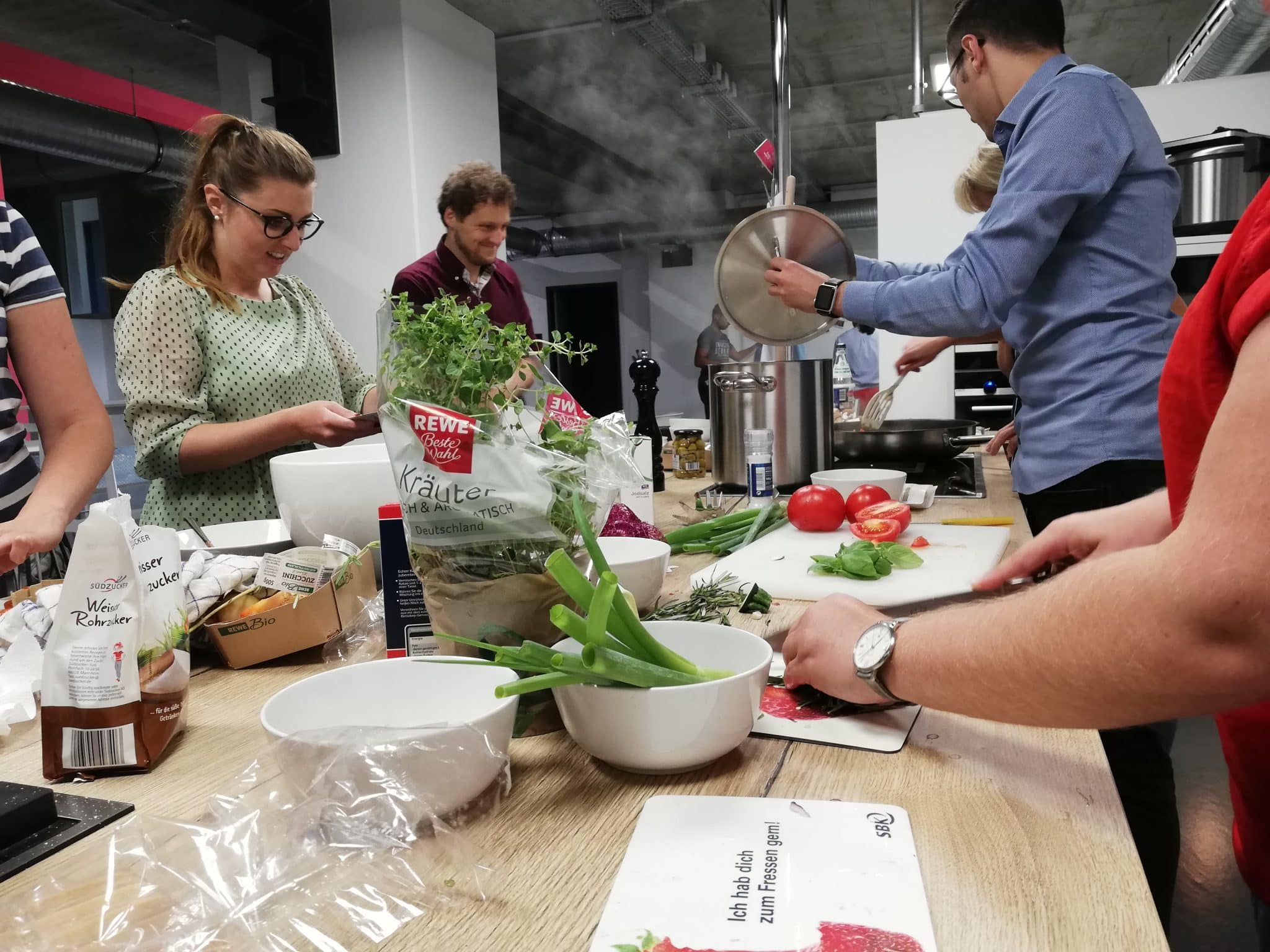 Menschen sind in einer Küche versammelt und bereiten gemeinsam Essen zu. Verschiedene Zutaten und Utensilien sind auf dem Tisch verteilt, darunter Kräuter, Gemüse und ein Schneidebrett. Eine Person rührt in einem großen Topf, während andere die Zutaten hacken oder ordnen. Die Atmosphäre ist geschäftig und kooperativ.