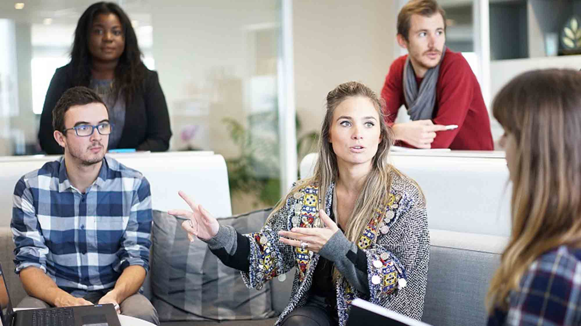 Eine Gruppe von fünf Personen diskutiert in einer modernen Büroumgebung. Zwei sitzen auf einer Couch, drei stehen um sie herum. Eine Frau gestikuliert beim Sprechen, die anderen hören aufmerksam zu. Laptops und Notizbücher sind zu sehen.