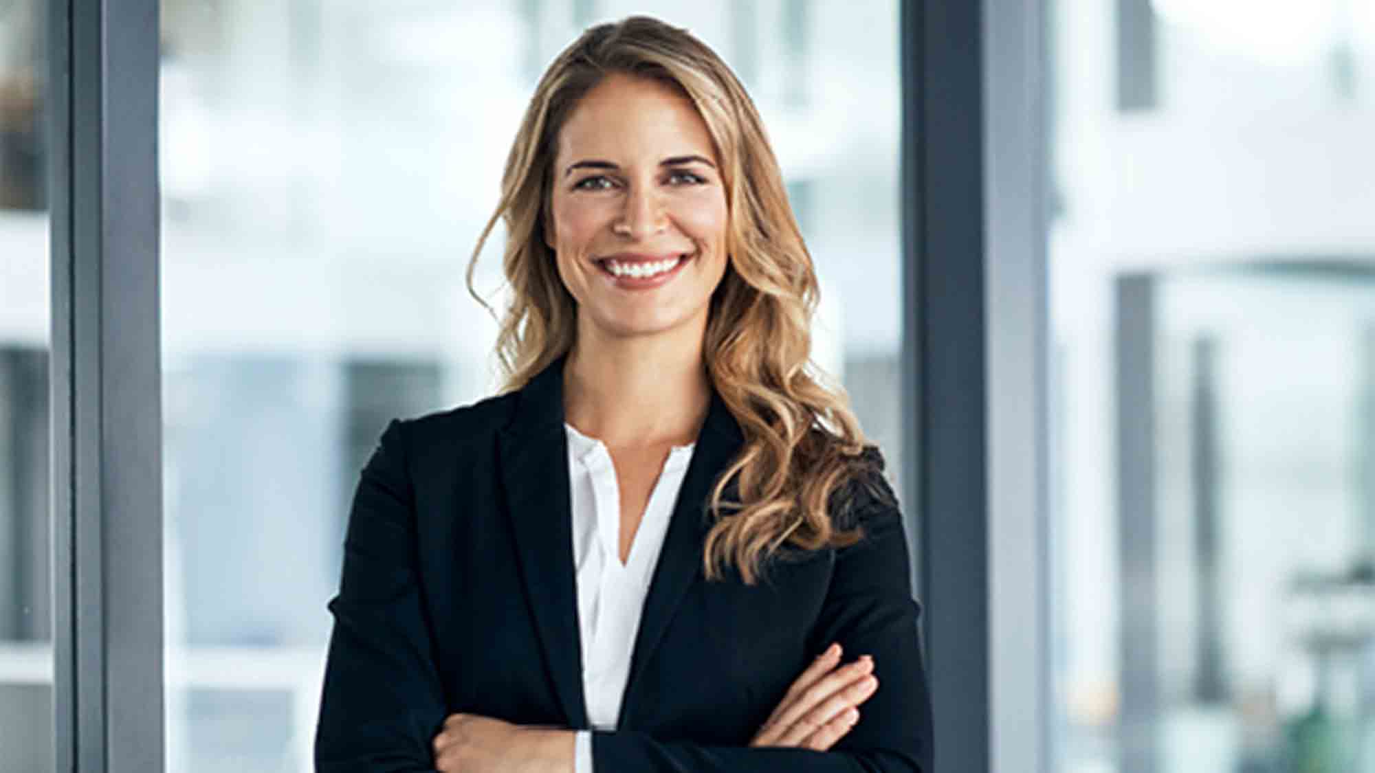 Eine selbstbewusste Frau mit langen blonden Haaren, einem schwarzen Blazer und einer weißen Bluse steht mit verschränkten Armen da und lächelt herzlich. Sie befindet sich in einem Büro mit großen Fenstern im Hintergrund.