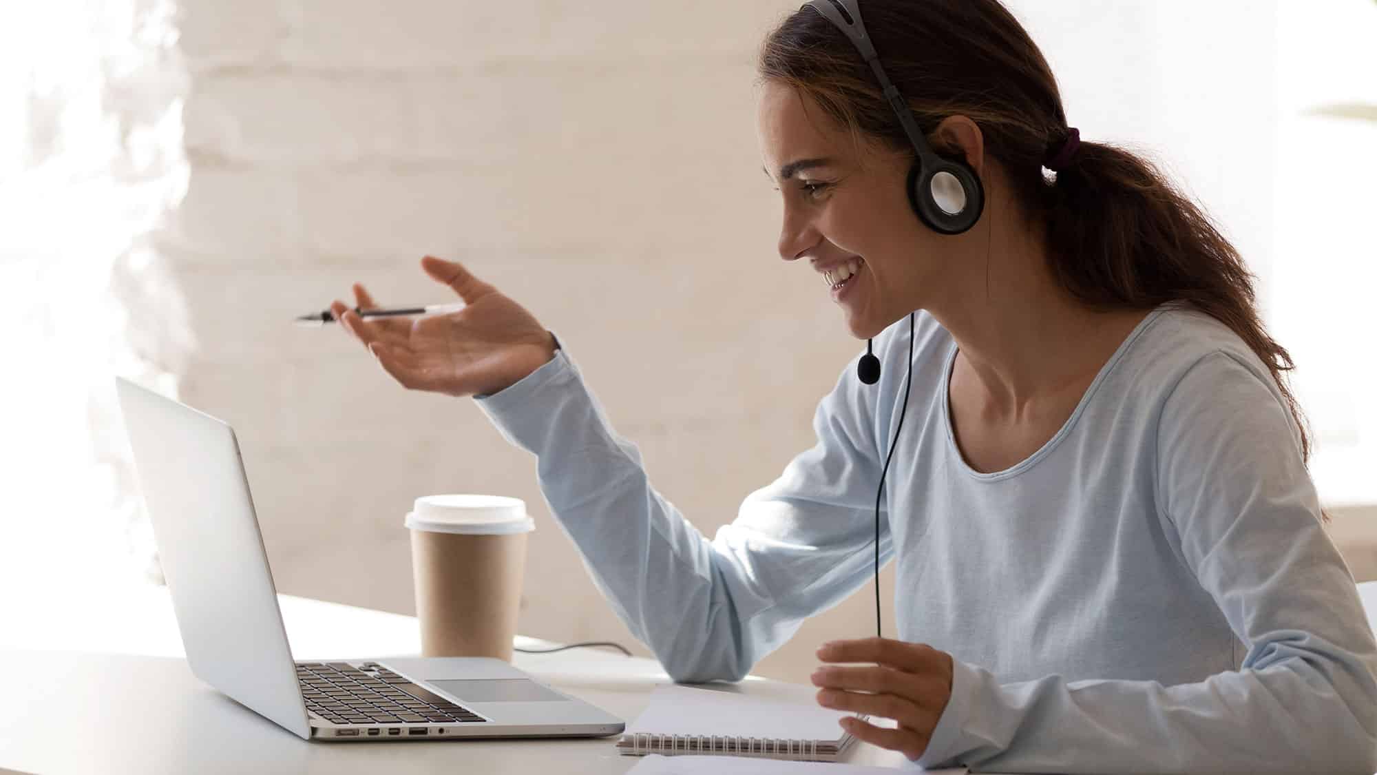 Eine Frau mit Headset sitzt mit einem Laptop an einem Schreibtisch, hält einen Stift in der Hand und lächelt. Ein Notizblock und eine Kaffeetasse liegen neben ihr, während sie an einem Videoanruf in einem hellen Raum mit einer weißen Ziegelwand im Hintergrund teilnimmt.